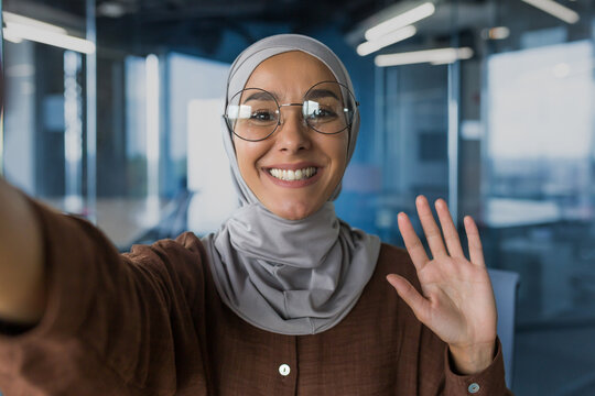 Young beautiful woman in hijab looking at smartphone camera, talking with friends on video call, muslim woman in glasses smiling and waving greeting gesture, working inside office modern building