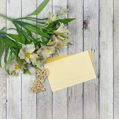 Elegant  paper blank- card in pastel sand color with envelope, white Alstroemeria  flowers ,two decorative hearts on the background of wooden table  in light colors. Greeting or invitation concept