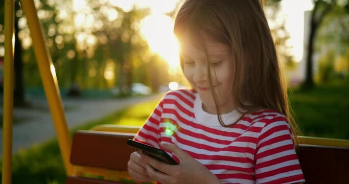 Portrait of little cheerful blonde girl uses a smart phone, ute caucasian child playing education game on her smartphone while sitting outdoors.