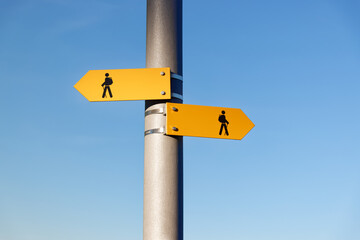 two yellow hiking trail signs with symbol person, backpack, hiking sticks, the signs point in different directions, in front of blue cloudless sky