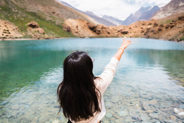 Young Indian female traveler pointing her finger towards Himalayan mountains while standing at clear blue water lake Deepak Tal, Himachal Pradesh, India. Travel and holiday concept. Summer in mountain