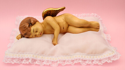 Figura de cerámica de ángel recostado sobre una cama blanca sobre fondo rosa claro. 
