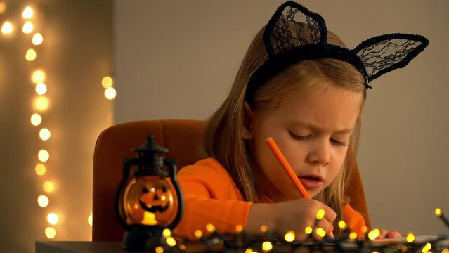 Close up of little girl sits in dark and makes decoration for holiday halloween. Child with pumpkin lamp on background of blurred lights of golden garland.