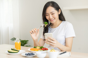 Obraz na płótnie Canvas Dieting, asian young woman eating, holding fork at broccoli, diet plan nutrition with fresh vegetables salad, enjoy meal while using smartphone. Nutritionist of healthy, nutrition of weight loss.