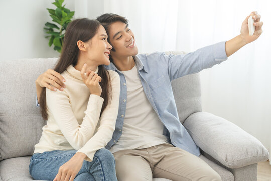Happy couple love at home, two asian young spending time, bonding to each other romantic on sofa in living room while man embrace woman using smartphone, mobile phone to selfie take a photo together.
