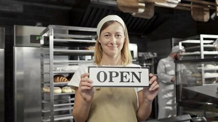 Portrait shot of Caucasian beautiful female baker in apron and hat standing in bakehouse in the morning and holding table Open. Bakery opening early concept. Good-looking woman chief in cafe.