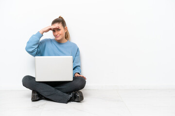 Young woman with a laptop sitting on the floor looking far away with hand to look something