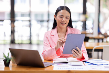 Asian Businesswoman working in the workspace, doing report analyzing financial figures on a graph in tablet and sitting on laptop computer 