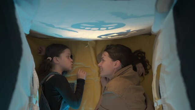 Happy funny young mom and cute little daughter girl lying on cozy dark tent decorated shadows pumpkin and bats in living room at home, family enjoying Halloween celebration, laughing, having fun