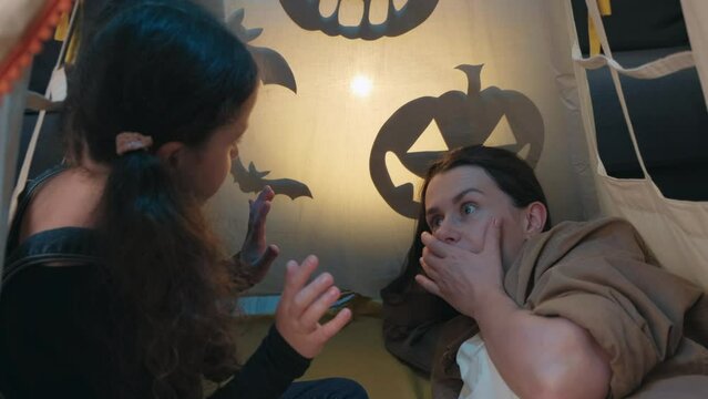 Close up of small daughter and young mother telling spooky stories using shadow play and paper bats and pumpkins. Mom and kid girl have fun, sitting on comfy tent at home. Puppet theater of shadows