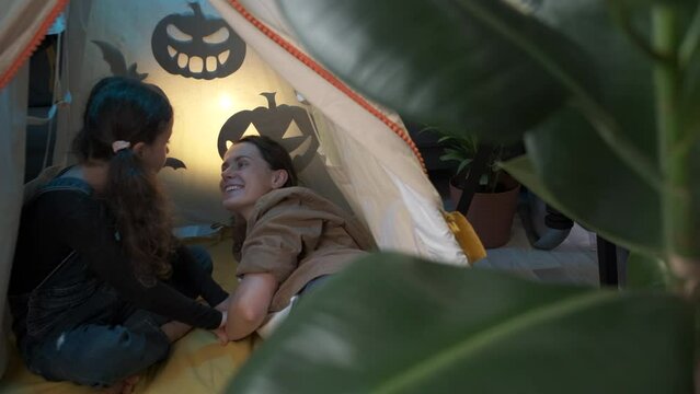 Cute fun little school daughter sitting in cozy tent with shadow bats and spooky pumpkins at home telling stories to loving young mother during Halloween celebration, family spending time together