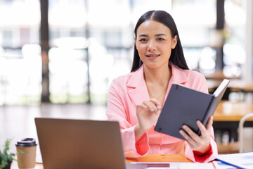 Obraz premium Asian Businesswoman holding notes and working on a laptop computer in the workplace, doing document planning analyzing the financial report, business plan investment, finance analysis concept.