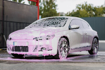 White sedan in pink soapy foam. Contactless self-service car wash concept. Personal car care...