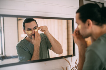 Spa day at home. Young man with patches under his eyes at the mirror at home.