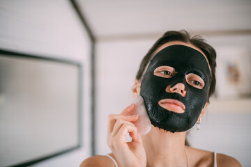 A beautiful brunette woman makes facial skin care procedures with a cleansing, moisturizing mask near the mirror in a bright room