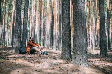Portrait of a young man sitting at the foot of a pine tree in a coniferous forest on a sunny day. A...