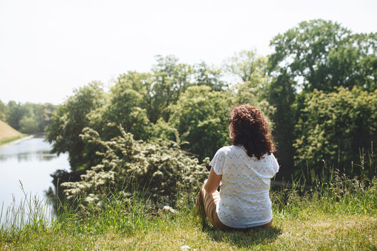 Back View Girl Traveler Sitting On Green Grass And Looking At Lake And Trees. Woman With Curly Hair. The Simple Pleasure Of Solitary Silence. Digital Detox. Copy Space For Text.