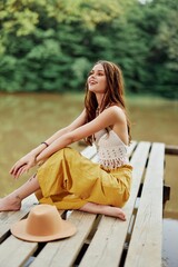 A hippie woman sits with her hat on a bridge by a lake with her hands up in the air on a nature trip and smiling happily in eco-clothing. Relaxed lifestyle