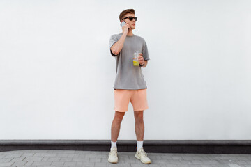 Fototapeta na wymiar Portrait of handsome confident stylish hipster man in sunglasses talking on the phone, standing guy near the concrete wall. Calm relaxing moment, summer vacation concept