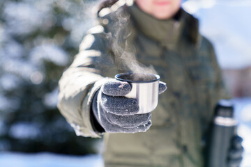 Man giving steamy coffee from thermos. Winter picnic in snowy wonderland. Traveler drinking tea and holding metal cup. Offer hot beverages. Lifestyle moment in nature. Close up of hands in gloves