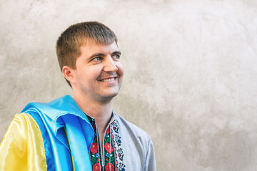 Close-up portrait of young man with blue and yellow flag of Ukraine. Man in traditional embroidered shirt against background of gray wall in city. Front view, Copy Space. Independence Day of Ukraine