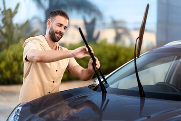 Handsome latin man replace windshield wipers on car standing outdoors. Car service concept  - obrazy, fototapety, plakaty