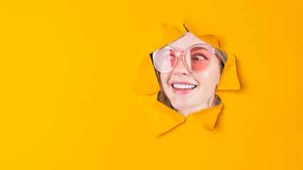 Beautiful face of young smiling woman with perfect teeth looking in paper torn hole in yellow background with copy space.