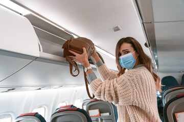 Woman traveler in mask putting luggage into overhead locker on airplane during boarding