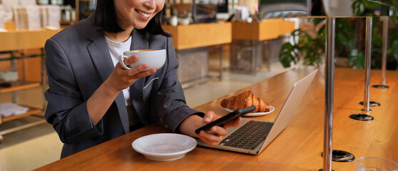 Young Asian business woman wearing suit drinking coffee using smartphone in cafe. Happy smiling female professional working holding mobile phone using smartphone texting messages on cellphone.