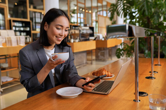 Young Asian business woman wearing suit drinking coffee using smartphone in cafe. Happy smiling female professional working holding mobile phone using smartphone texting messages on cellphone.