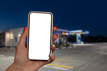 Woman holds a close-up of a smartphone with a white screen in his hands against the backdrop on a gas station. Technology mockup for apps and websites.