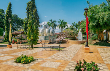 Fototapeta premium A nice and relaxed park with a wooden bridge over a water fountain, Traditional park of Nagarote, Nicaragua. View of a calm park with a small wooden bridge on a sunny day. Nagarote central park