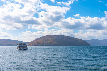 Lake Towada Sightseeing Cruises. Beautiful view, clear blue sky, white cloud, cruise ship in sunny day with autumn foliage season background. Aomori, Japan. Text in Japanese on ship 