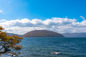 Beautiful autumn foliage scenery landscapes. Fall is full of magnificent colors. View from shore of Lake Towada, clear blue sky and water, white cloud, sunny day background. Aomori Prefecture, Japan