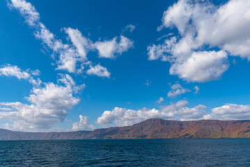 Beautiful autumn foliage scenery landscapes. Fall is full of magnificent colors. View from Lake Towada sightseeing Cruise ship. Clear blue sky, water, white cloud, sunny day background. Aomori, Japan