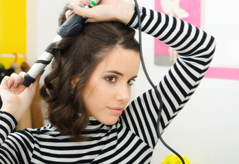 Beautiful young woman curling her hair with hair curler in front of the room mirror