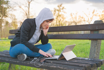 Young woman in hoodie sitting on the bench in the park and using laptop and smart phone in autumn