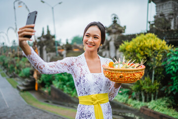 attractive balinese young woman taking her selfie photo with mobile phone while wearing traditional kebaya clothes in bali