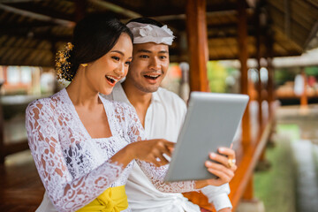 balinese man and woman with traditional clothes using tablet pc while sitting together
