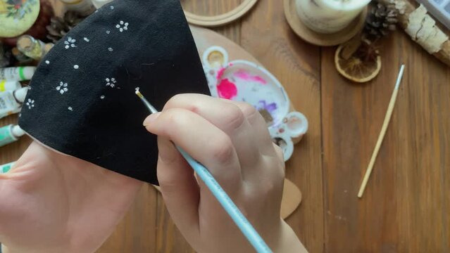 Woman draw flowers on medical cloth mask with brush and paint. Design of accessories in coronavirus pandemic. Wooden table with palette, acrylic in tubes, candle and other objects. Quarantined hobby.
