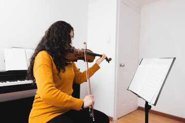 Brunette woman wearing a yellow sweater, and playing violin by reading sheet music.