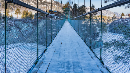 A suspended pedestrian bridge with a lattice railing leads to a secluded monastery on the island. The snow-covered plank surface of the path. A frozen river is visible, a sunlit mountain range. Altai