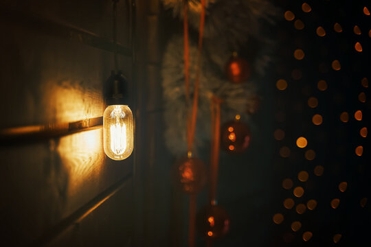 Vintage type bulb and fir branches with red balls and ribbons on wooden wall. Christmas decorations. Copy space for New Year congratulations. Blurred warm garland lights on background. Party mood.
