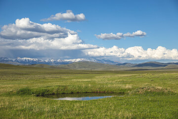 Fototapeta na wymiar Pond in green plain, with snowy mountains and blue sky with clouds