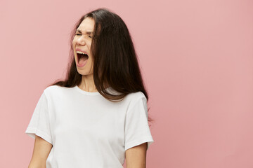 Fototapeta premium emotional, angry, woman with dark hair color stands in a white t-shirt on a pink background and screams loudly. Horizontal photo with blank space for advertising text