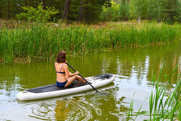 A woman drives on the Sup Board through a narrow canal surrounded by dense grass. Active weekend vacations wild nature outdoor. A woman is sitting with her legs stretched out.