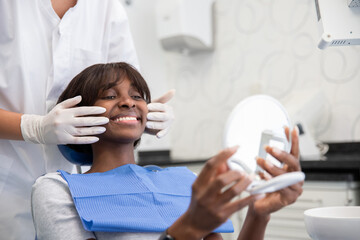 Smiling woman sitting in dentists chair and looking at teeth. Young African American female patient holding mirror and examining teeth with dentist standing behind.Oral hygiene and dental care concept