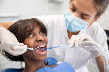 Female dental hygienist using dental hook and suction during cleaning teeth procedure. Young African American woman visiting stomatology clinic. Oral hygiene and dental care concept - obrazy, fototapety, plakaty