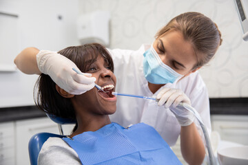 Young female dentist treating teeth of African American woman in stomatology clinic. Dental hygienist cleaning teeth of young patient using hook. Oral hygiene and dental care concept - obrazy, fototapety, plakaty