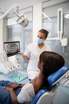 Confident female dentist showing x-ray picture of jaw to patient. Young African American woman sitting in dentist chair. Visiting dentist concept